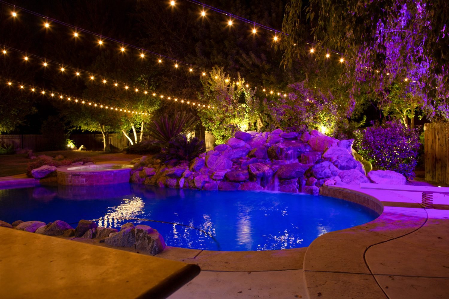 Warm string lights above a Bakersfield pool with a color-lit rock waterfall at night